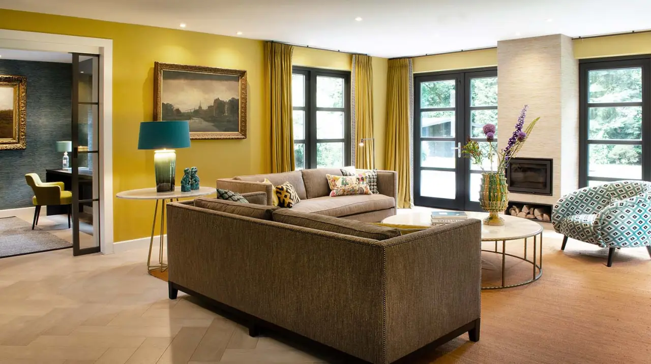 Woonkamer met gele wanden en gordijnen, bruine bank en een fauteuil met dessin
